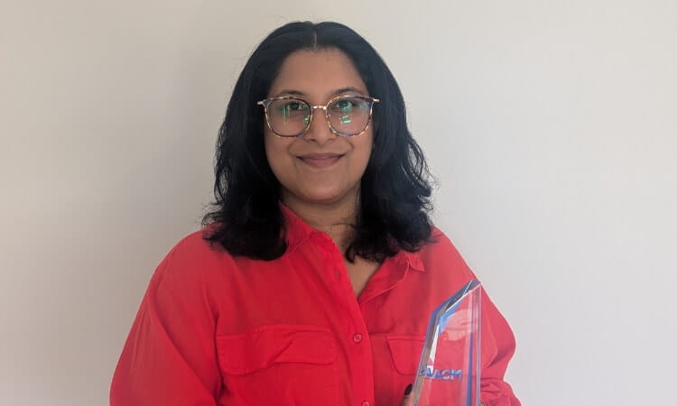 A person with shoulder-length black hair, wearing glasses and a red shirt, holds a transparent award in front of a plain white background.