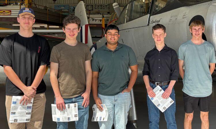 Five young men stand in a hangar holding papers, posing in front of a silver aircraft.