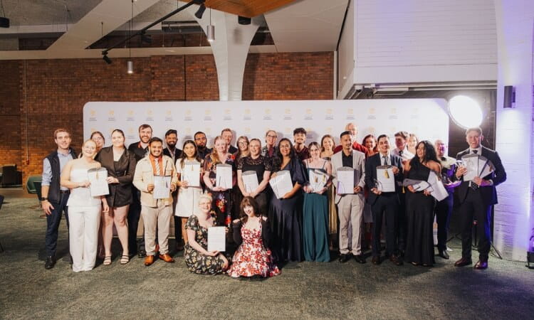 A large group of people dressed formally stand indoors, posing and holding certificates in front of a branded backdrop at an event.
