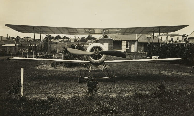 A vintage biplane with a large propeller is parked on grass in a residential area, with houses, fences, and trees in the background.