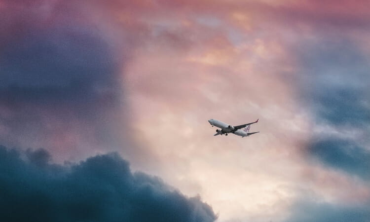 A commercial airplane flies through a dramatic sky with dark clouds and a colorful sunset or sunrise in the background.