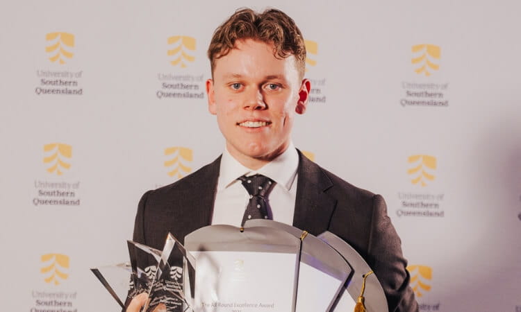 A young man in a suit holds multiple awards and certificates, standing in front of a University of Southern Queensland backdrop.
