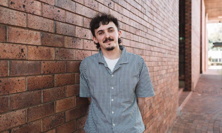 A person with short dark hair and a mustache stands against a brick wall, wearing a short-sleeved, checkered button-up shirt.