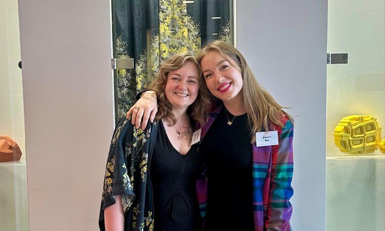 Two women stand indoors, smiling at the camera with one arm around each other. Both wear name tags and formal attire. Geometric art pieces are visible in the background.
