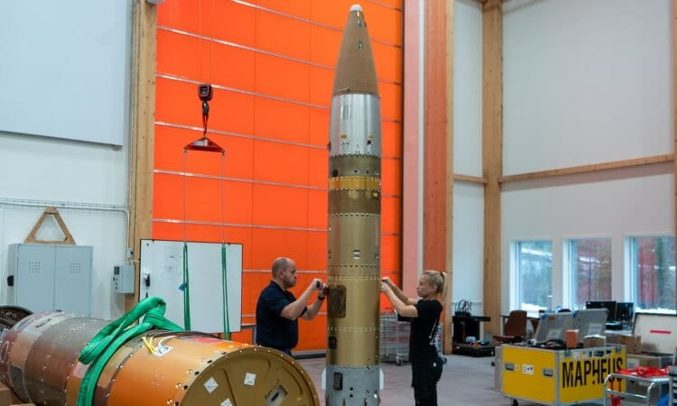 Two people work on assembling a large missile or rocket inside an industrial facility with orange walls and equipment nearby.