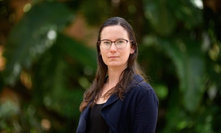 A person with long brown hair and glasses wearing a dark jacket stands outdoors in front of green foliage.