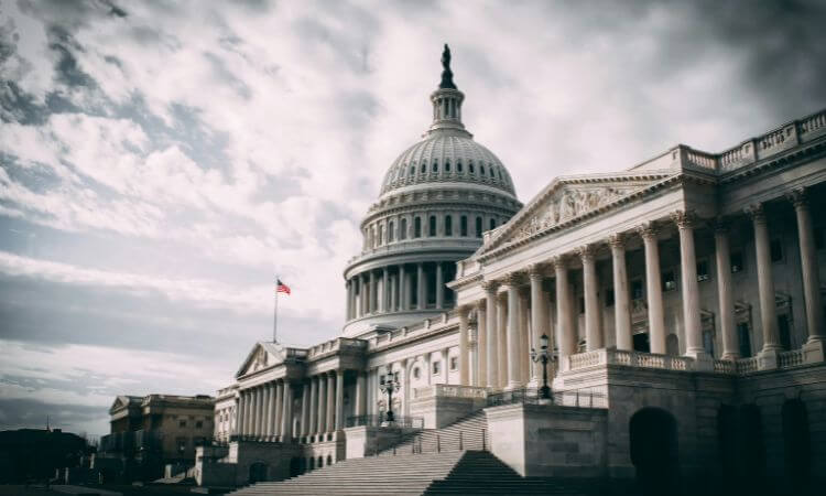 The United States Capitol building with its dome and columns under a cloudy sky, and an American flag flying outside.