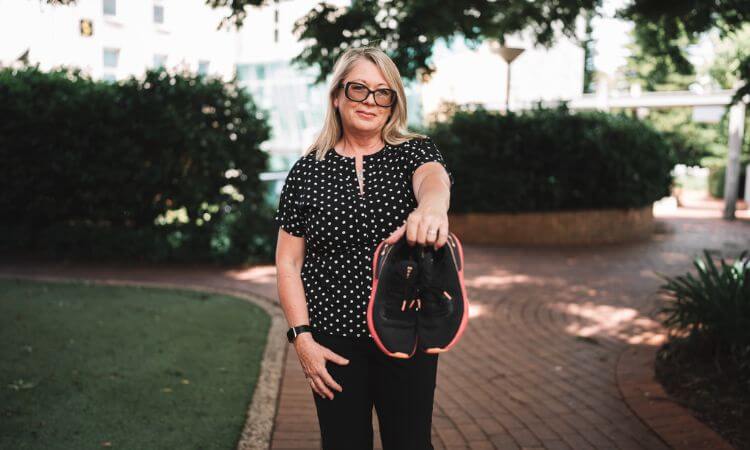 A woman with blonde hair and glasses stands outdoors on a path, holding a pair of black athletic shoes with pink accents toward the camera.