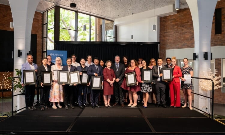 A group of people stand on a stage, many holding framed certificates, posing for a formal photo at an indoor award ceremony.