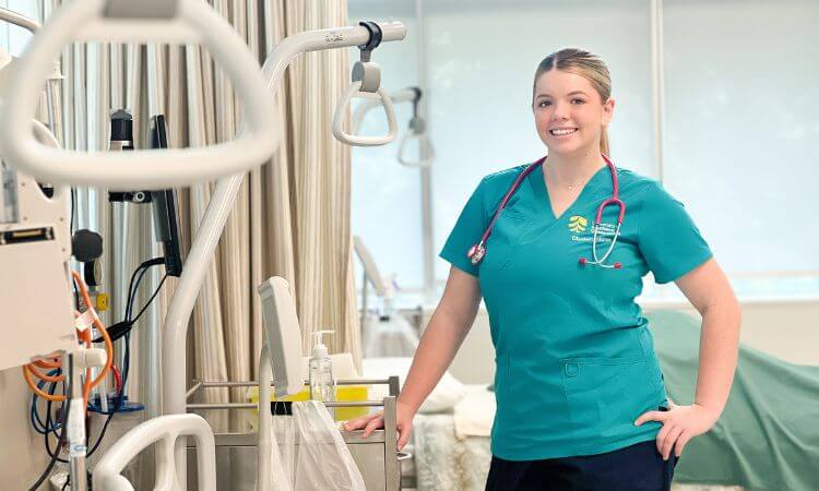 A nurse in teal scrubs with a stethoscope stands in a medical room beside healthcare equipment, smiling at the camera.