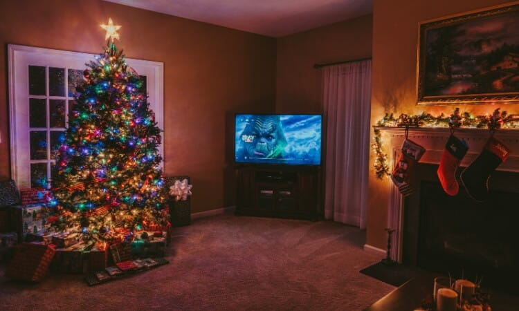 A living room decorated for Christmas with a lit tree, presents underneath, stockings on the fireplace, and a TV displaying a movie with a monster.