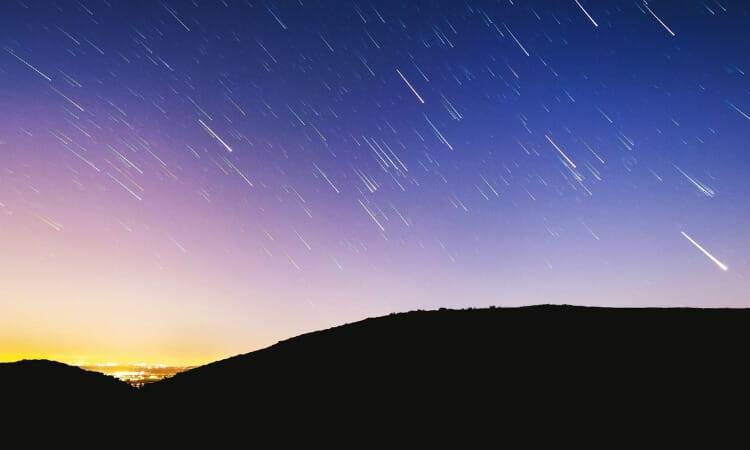 Long-exposure photo of the night sky showing star trails above a dark silhouetted hill with a city’s lights visible on the horizon.