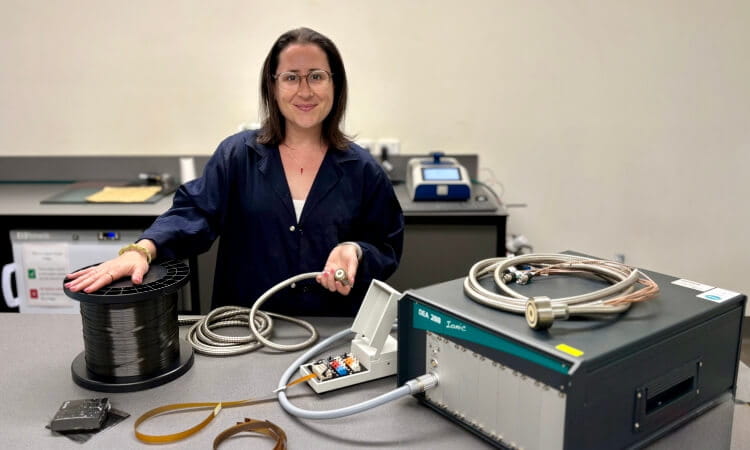A woman in a lab coat stands at a workbench with scientific equipment, holding a cable beside a large machine and a spool of wire.