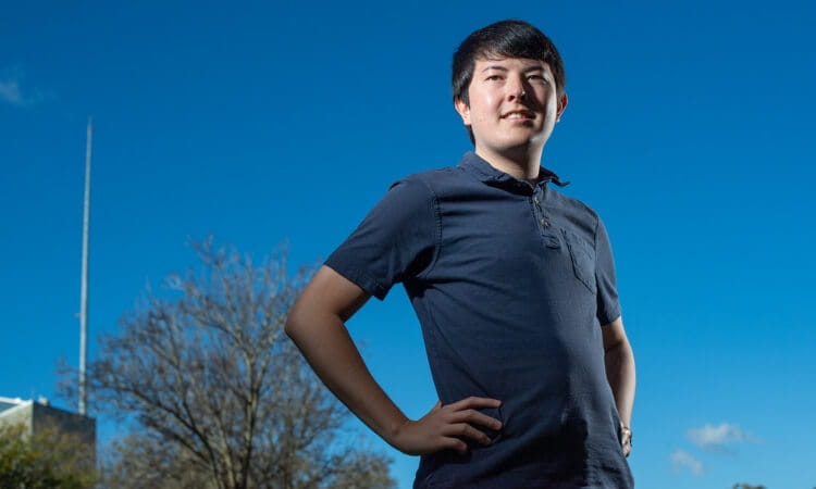 A person stands outdoors with hands on hips, wearing a navy shirt, under a clear blue sky with some trees and a structure in the background.