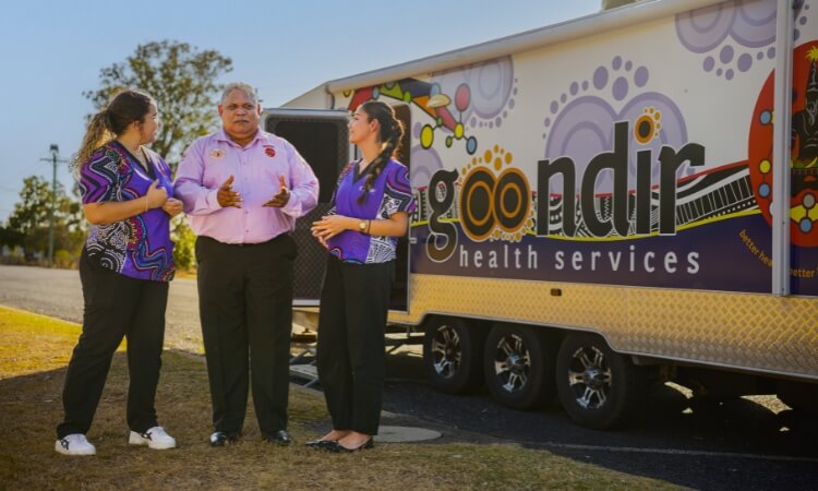 Three people stand and talk in front of a Gooldir Health Services mobile unit.