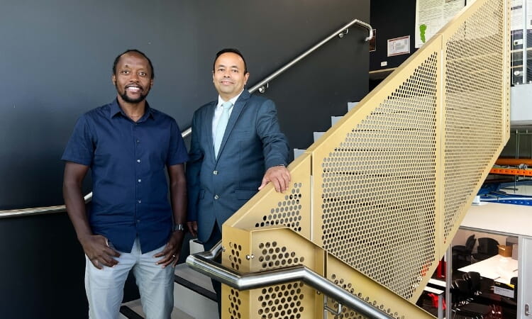 Two men stand by a staircase indoors, one in a navy shirt and khaki pants, the other in a blue suit, posing for a photo against a black wall background.