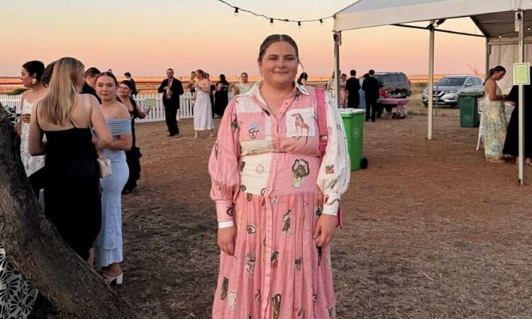 A woman in a pink patterned dress stands outdoors at an event, with people socializing in the background near a white tent at sunset.
