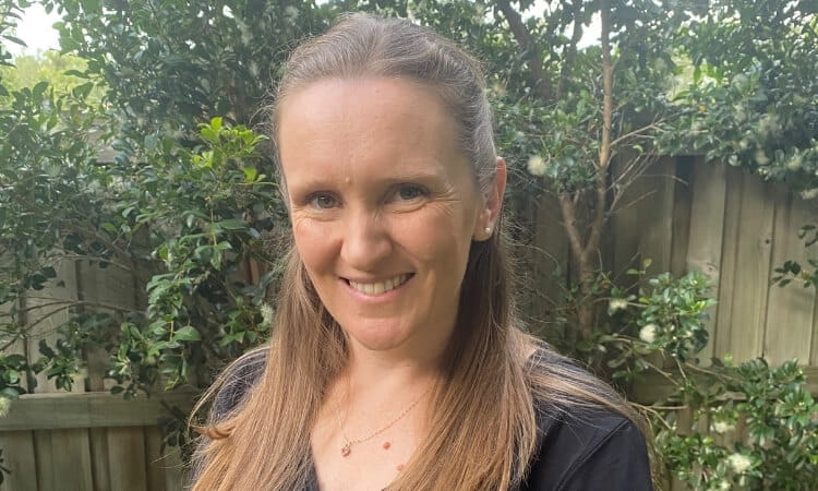A woman with long light brown hair smiles at the camera while standing in a garden with green trees and a wooden fence in the background.