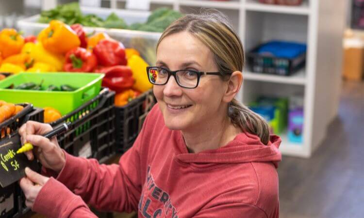 A woman wearing glasses and a red hoodie labels produce at a market, with bell peppers and other vegetables arranged behind her.