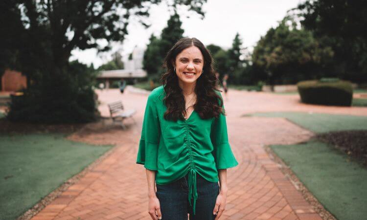 A woman with long brown hair, wearing a green blouse and jeans, stands smiling on a brick pathway in an outdoor park setting.