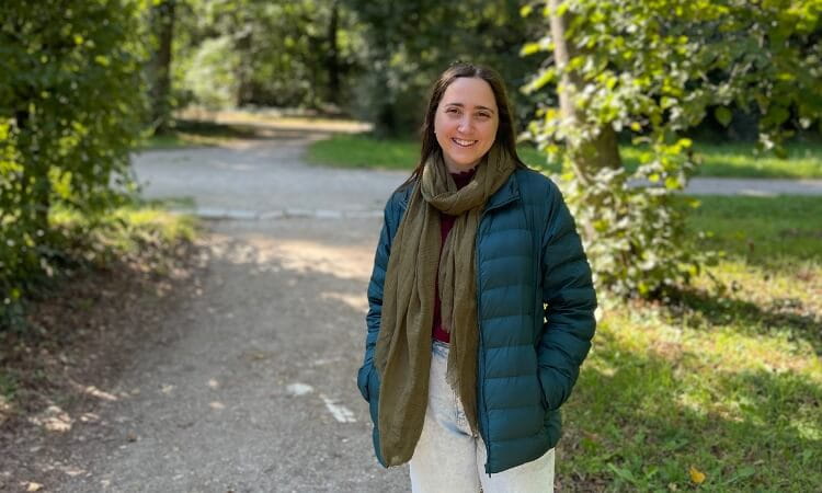 A woman wearing a green jacket and scarf stands on a sunlit park path with trees and greenery in the background.