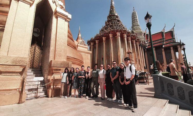 A group of people stands in front of ornate golden temple buildings under a clear blue sky.