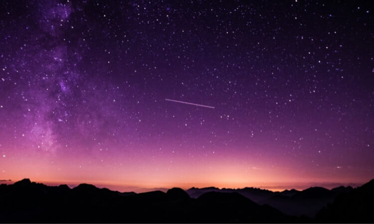 A starry night sky with a faint Milky Way, a shooting star, and purple hues above silhouetted mountain peaks.