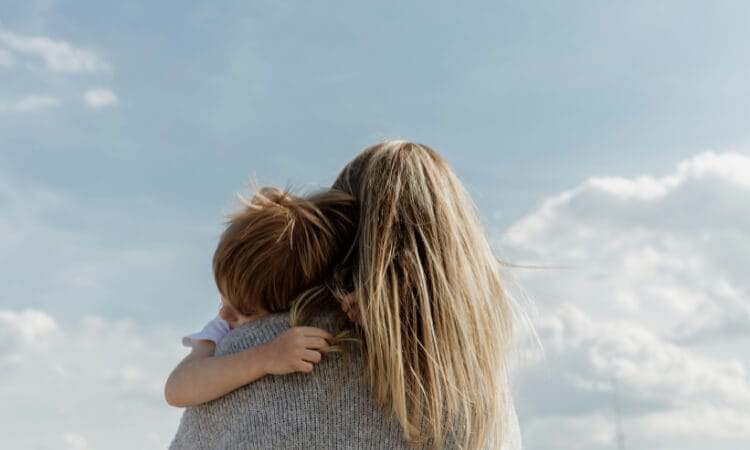 A woman with long blonde hair holds a small child, both facing away from the camera, under a partly cloudy sky.