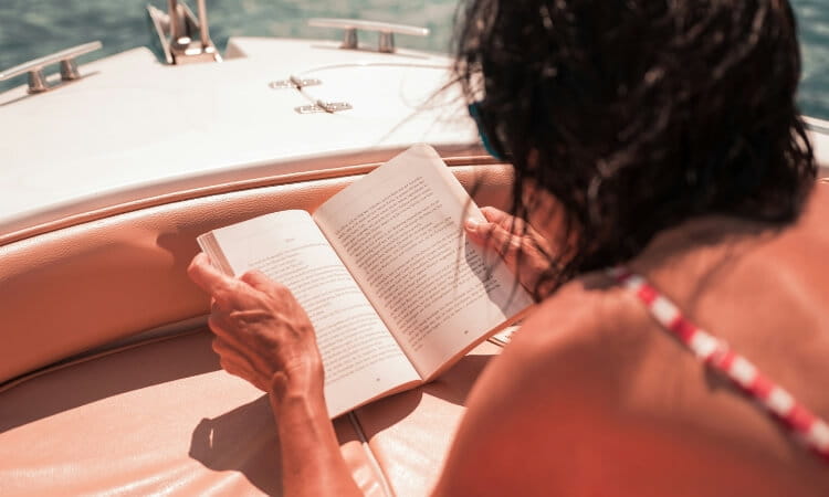 Person in a swimsuit lies on a boat deck reading a book, with sunlight illuminating the scene and water visible in the background.
