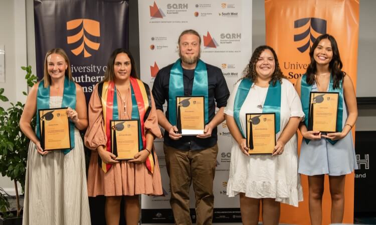 Five people stand in a row holding award plaques and wearing teal sashes, posing in front of University of Southern Queensland banners.
