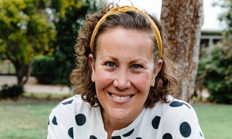 A person with short curly hair, wearing a yellow headband and a white shirt with black polka dots, smiles outdoors in a park setting.