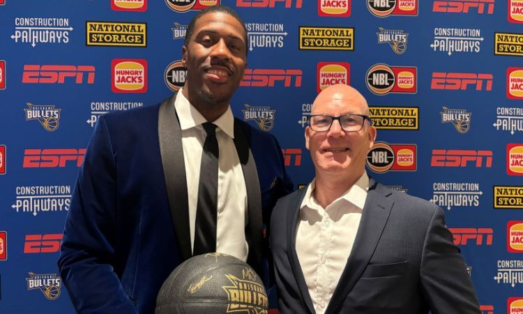 Two men in suits pose together at an event; one holds a black basketball. The backdrop displays various sponsor logos including ESPN and National Storage.