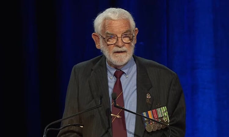 Elderly man in a suit and tie, wearing glasses and military medals, speaks at a podium against a blue background.