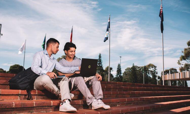 Two people sit on outdoor brick steps with a laptop, surrounded by flagpoles and trees under a blue sky.
