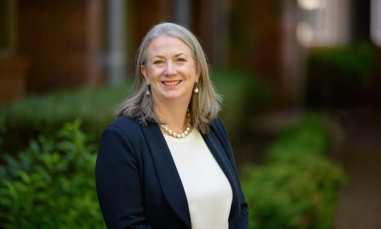 A woman with gray hair wearing a navy blazer, white top, and pearl necklace stands outside, smiling, with green bushes and buildings in the background.
