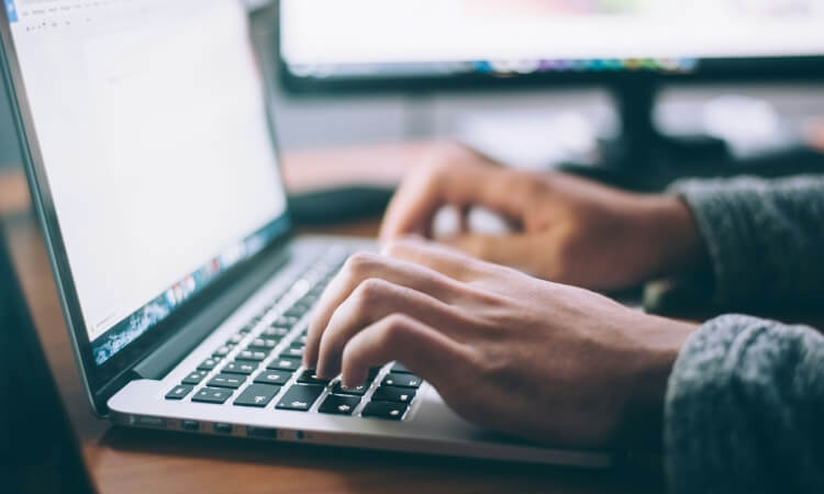 A person types on a laptop keyboard at a desk with a computer monitor in the background.