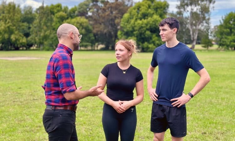 A man in a plaid shirt talks to a woman and a man in athletic wear standing together on a grassy field with trees in the background.