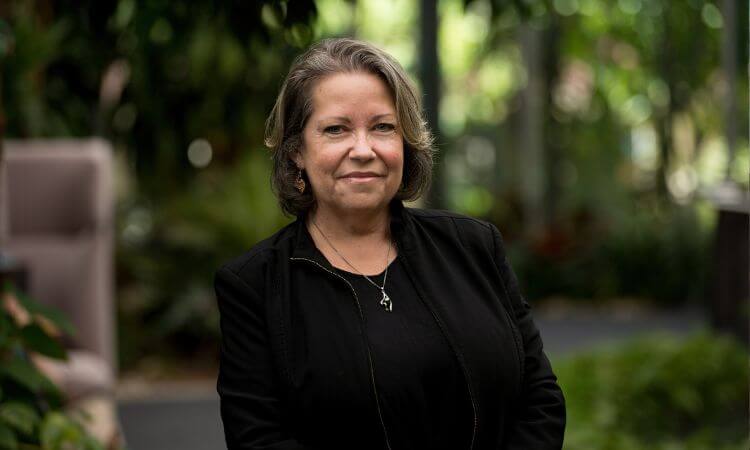 A woman with shoulder-length brown hair wearing a black jacket stands outdoors in a garden setting, looking at the camera and smiling slightly.