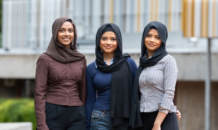 Three young women wearing hijabs stand close together outdoors, smiling at the camera.