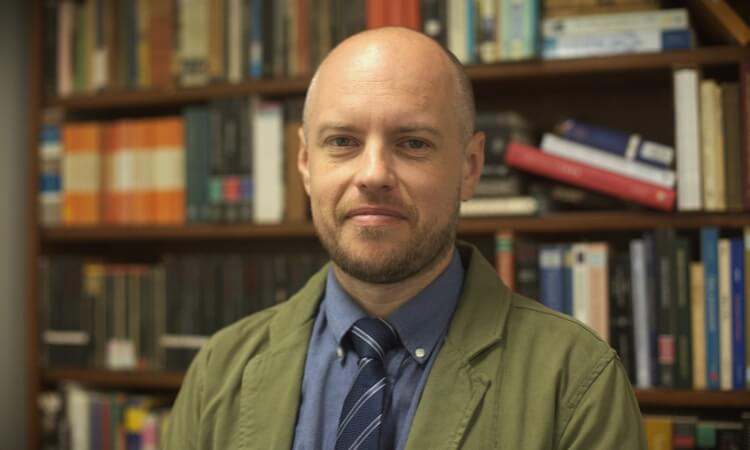 A man with a shaved head and beard, wearing a green jacket and blue shirt with a tie, stands in front of bookshelves filled with books.