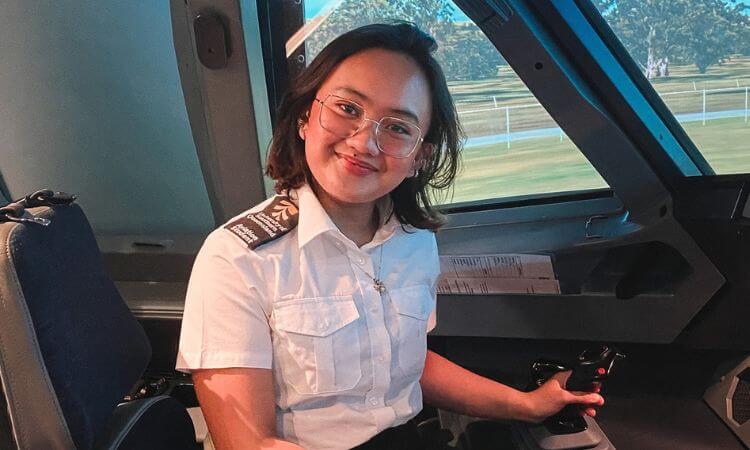 A woman wearing a pilot uniform and glasses sits in an aircraft cockpit, smiling at the camera with her hand on the flight controls.