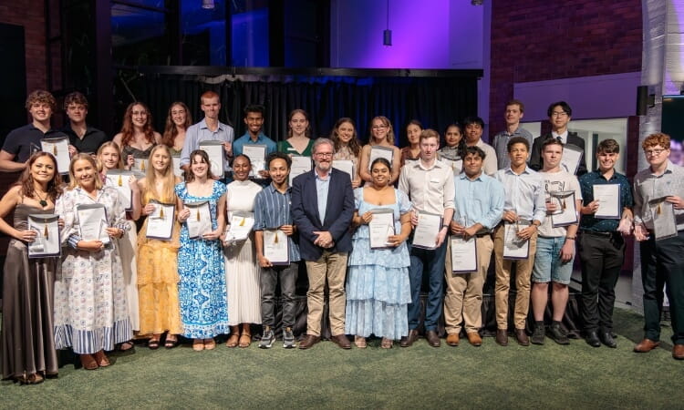A group of young adults and two older men stand in rows, dressed formally, holding certificates, and posing for a group photo at an indoor event.