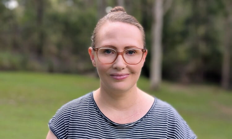 A woman wearing glasses and a striped shirt stands outdoors with greenery and trees in the blurred background.