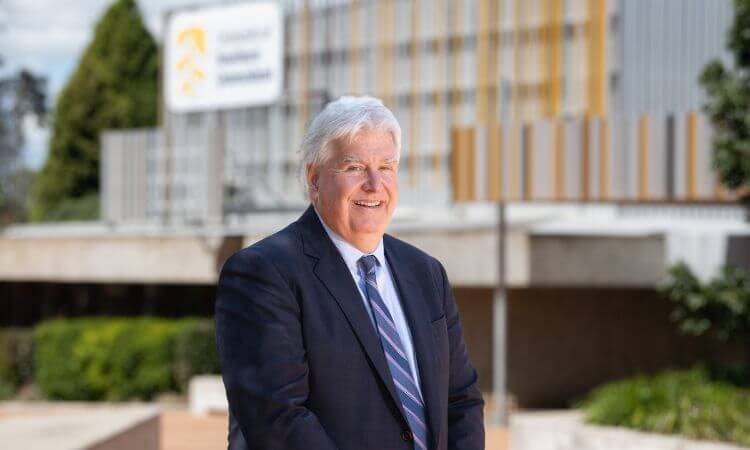 A man in a suit and tie stands outside a modern building with a blurred sign and landscaped greenery in the background.