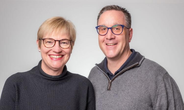 Two adults with short hair and glasses, wearing dark sweaters, stand side by side smiling in front of a plain light grey background.
