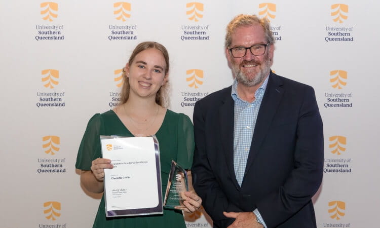 Two people stand in front of a University of Southern Queensland backdrop; the woman holds a certificate and a glass award, both are smiling.