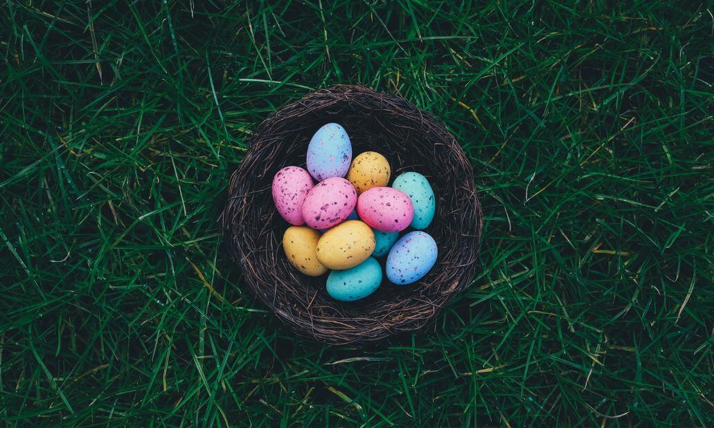 A bird’s nest filled with colorful speckled eggs, including pink, yellow, blue, and purple, sits on green grass.