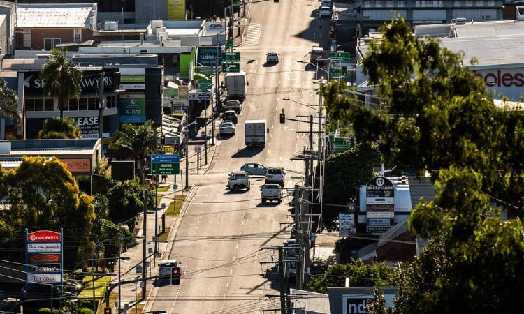 A city street with cars at an intersection, surrounded by commercial buildings, billboards, and trees in the foreground.