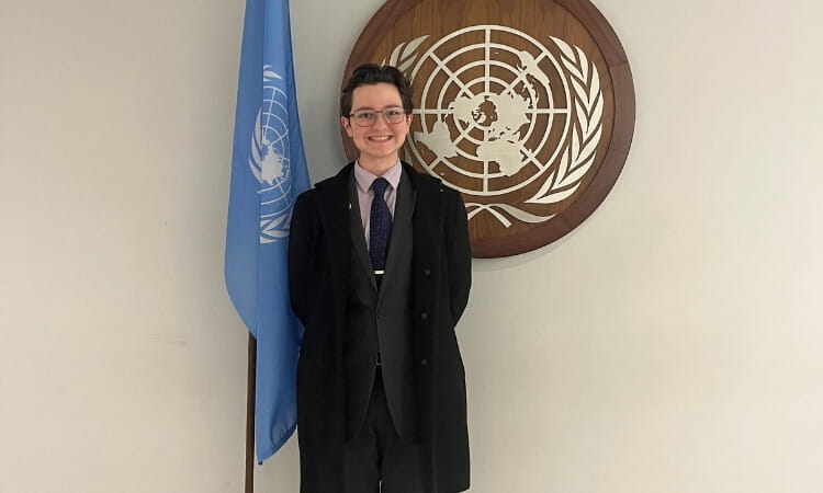 A person in formal attire stands in front of a United Nations emblem and flag, smiling at the camera.