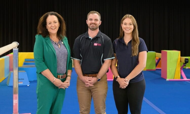 Three adults stand side by side in a brightly colored indoor gym with gymnastic equipment in the background.